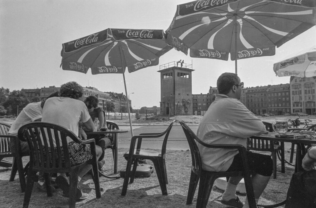 Schwarzweißfoto von Menschen, die an Plastiktischen unter Sonnenschirmen sitzen; im Hintergrund Gebäude und ein Turm